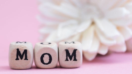 "MOM" écrit sûr des dés dans un fond rose et en arrière plan une fleur blanche.